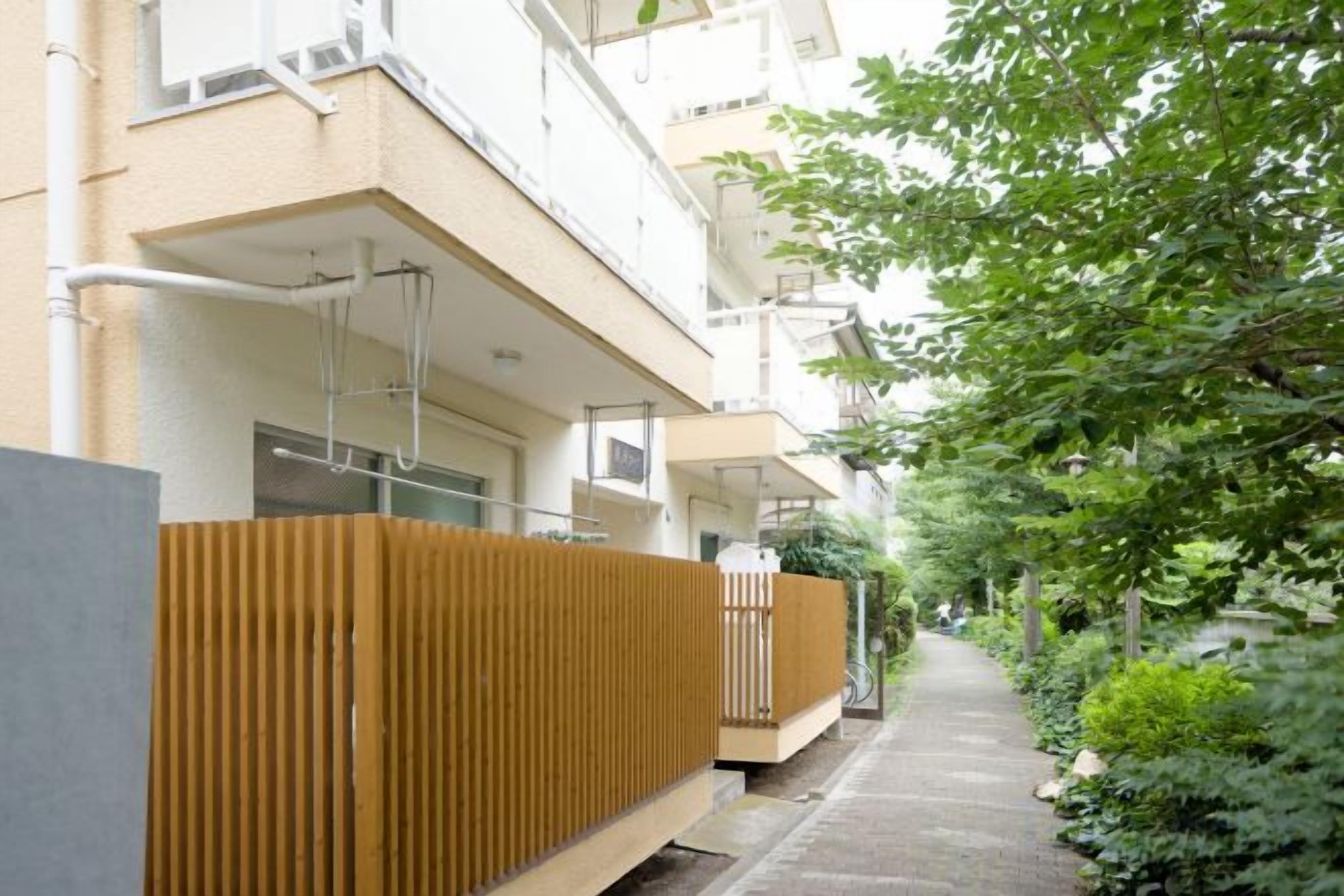 A narrow pathway flanked by apartment buildings with balconies, a wooden fence, and greenery.