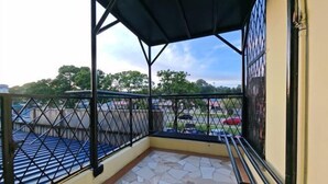Uma varanda coberta com piso de azulejos, guarda-corpo de metal e vista para um estacionamento e árvores.