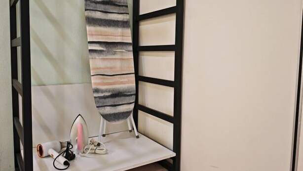 A room with a white ironing board against a wall, a small shelf with an iron, and a pair of shoes. - Kepayan Inn Kinabalu City (Penampang)