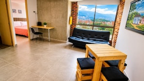 A room with a wooden table, black cushioned bench, a black sofa, a small desk with a chair, and a view of a cityscape through a large window.