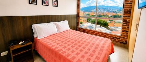 Habitación de hotel con una cama, una mesita de noche con teléfono y un ventanal que ofrece vistas a la ciudad.
