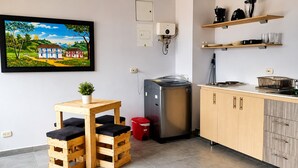 Een kleine keuken met een koelkast, een spoelbak en houten kastjes. Er staat een tafeltje met een bloempot en een rode prullenbak.