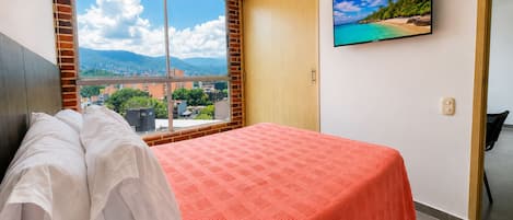 Habitación de hotel con cama, un ventanal con vistas a la ciudad, televisor en la pared y puerta que da a otra habitación.