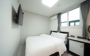A hotel room with a bed, a desk, a flat-screen TV, an air conditioner, and a window with curtains.