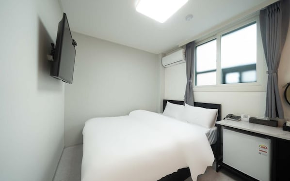 A hotel room with a bed, a desk with a telephone, an air conditioning unit, and a window with curtains. - Doksan White (Seoul)
