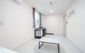 A small, clean room with a bed, a desk, and a chair. There is a window with curtains, a radiator, and a door with a coat hanging on it. - Doksan White (Seoul)