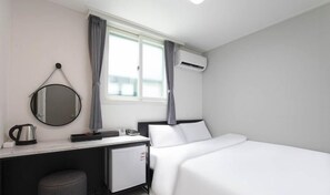 A hotel room with a bed, a desk with a mirror, a kettle, a small fridge, and a window with curtains. - Doksan White (Seoul)