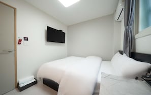 A hotel room with a bed, a flat-screen TV mounted on the wall, a window with curtains, and a small bedside table.