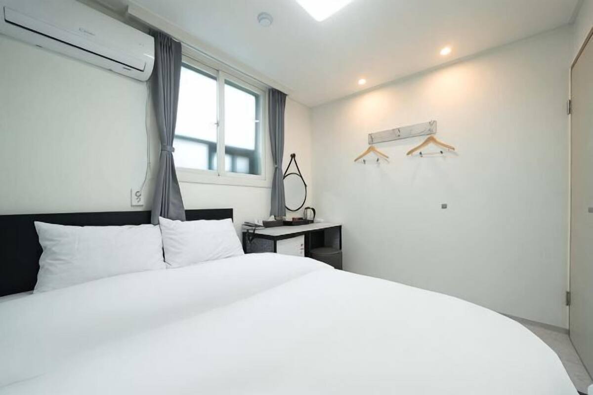 A hotel room with a large bed, a desk with a lamp, a window with curtains, and a coat rack.