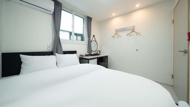 A hotel room with a large bed, a desk with a chair, a hanging coat rack, and a window with curtains. - Doksan White (Seoul)