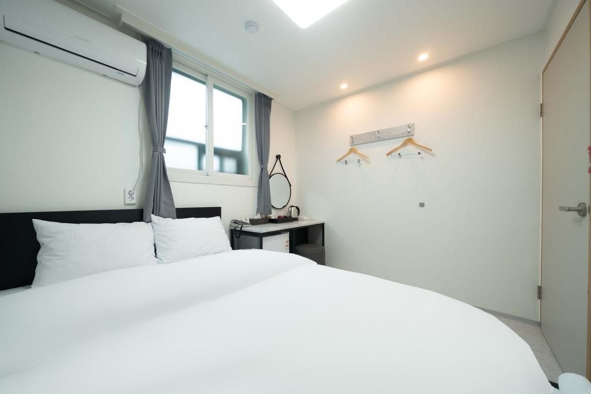 A hotel room with a large bed, a desk with a chair, a hanging coat rack, and a window with curtains.
