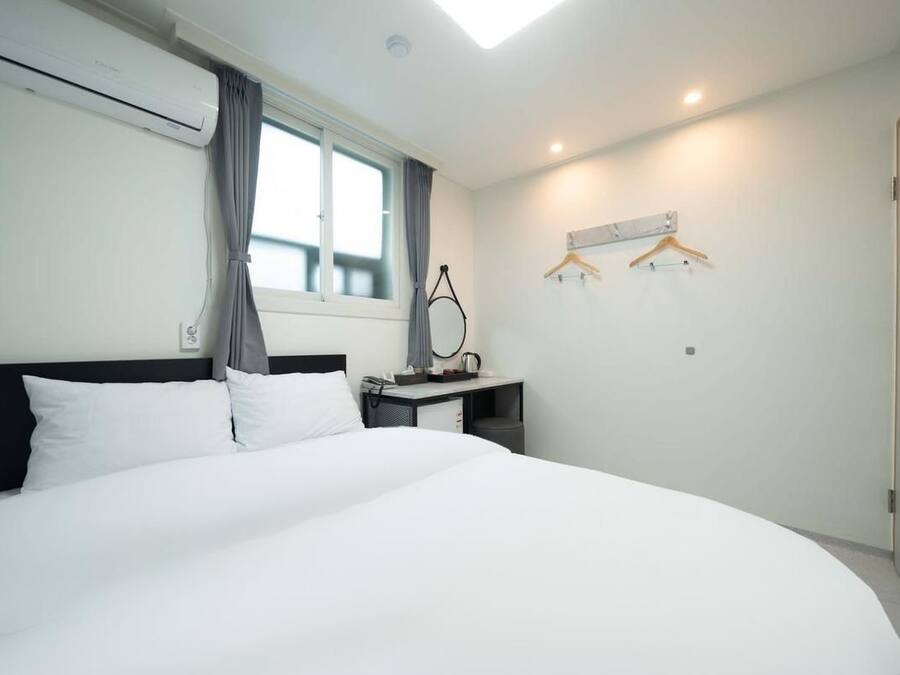 A hotel room with a large bed, a desk with a chair, a hanging coat rack, and a window with curtains.