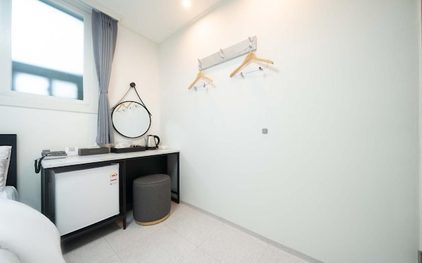 Un dormitorio moderno con cama, escritorio con computadora, un refrigerador pequeño, un cesto de basura y una ventana con cortinas.