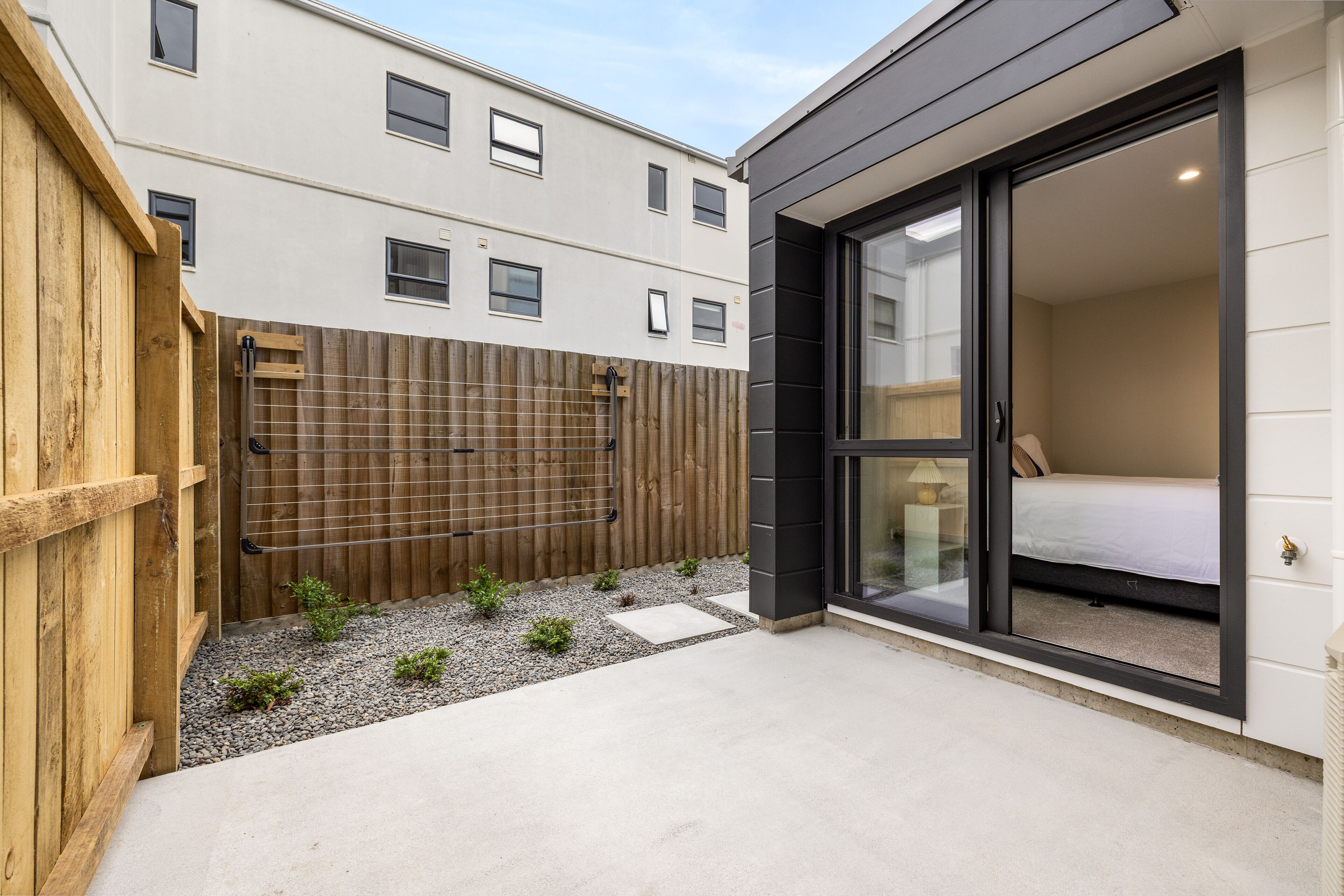 A modern building with a wooden fence, a small garden area with gravel and plants, and a bedroom with a bed and a window.