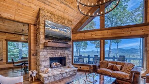 A cozy cabin interior with a stone fireplace, wooden furniture, and large windows offering scenic views. - New! Mtn Views, pool table, fire pit (Blue Ridge)