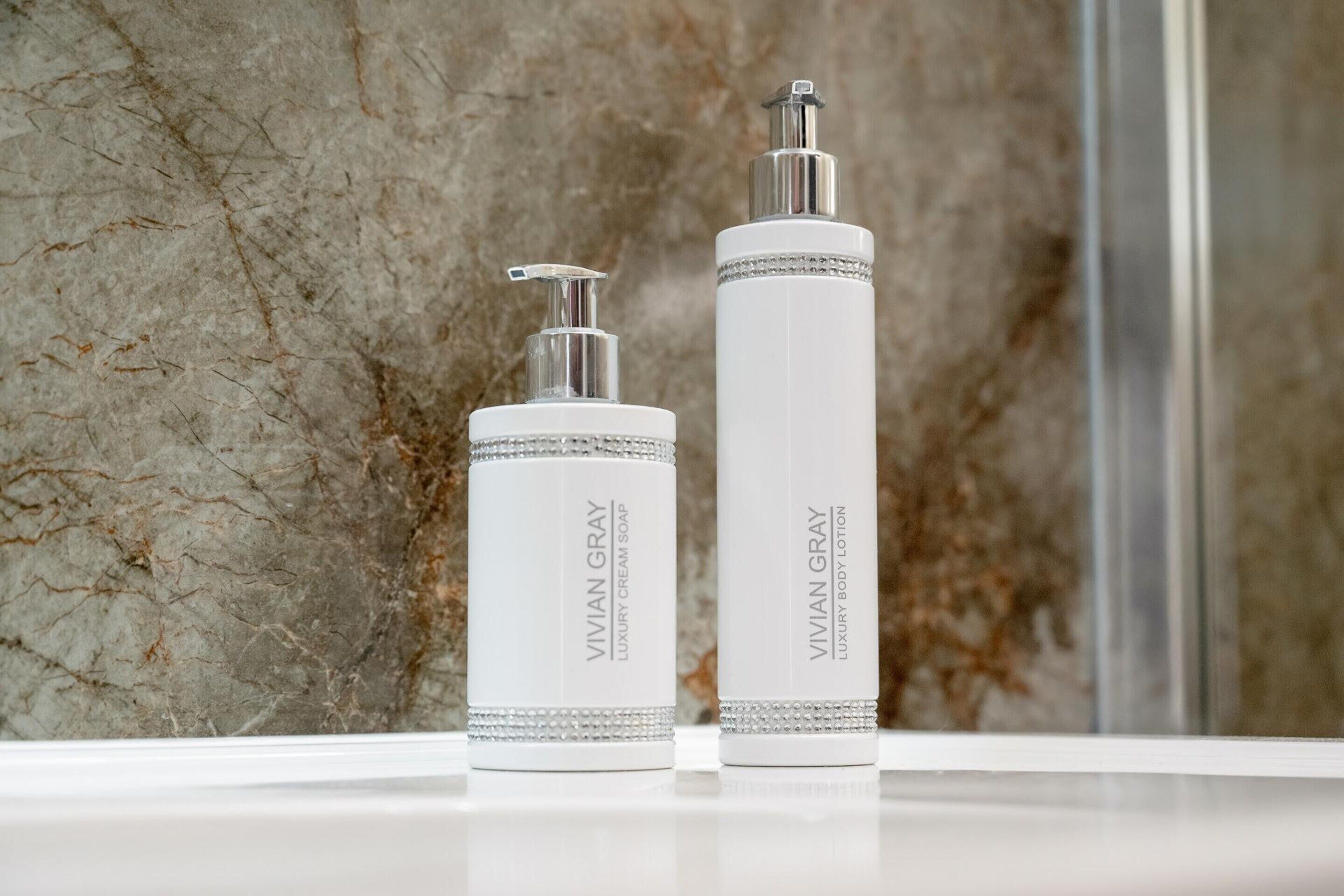 Two bottles of VIVIAN GRAY luxury body lotion and cream soap on a white surface against a marble wall.