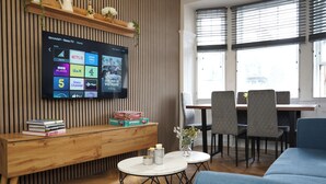Uma sala de estar moderna com uma TV de tela plana fixada em uma parede de madeira, um banco de madeira com livros, uma mesinha com objetos decorativos e um sofá azul.