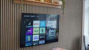 Televisão de tela plana fixada na parede, com design de painel de madeira, exibindo a interface de um serviço de streaming com vários canais.