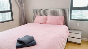 A neatly made bed with pink bedding and a folded gray towel, a striped headboard, and a small bedside table with drawers. - Thao Dien Masteri Riverside - Saigon Home (Ho Chi Minh City)