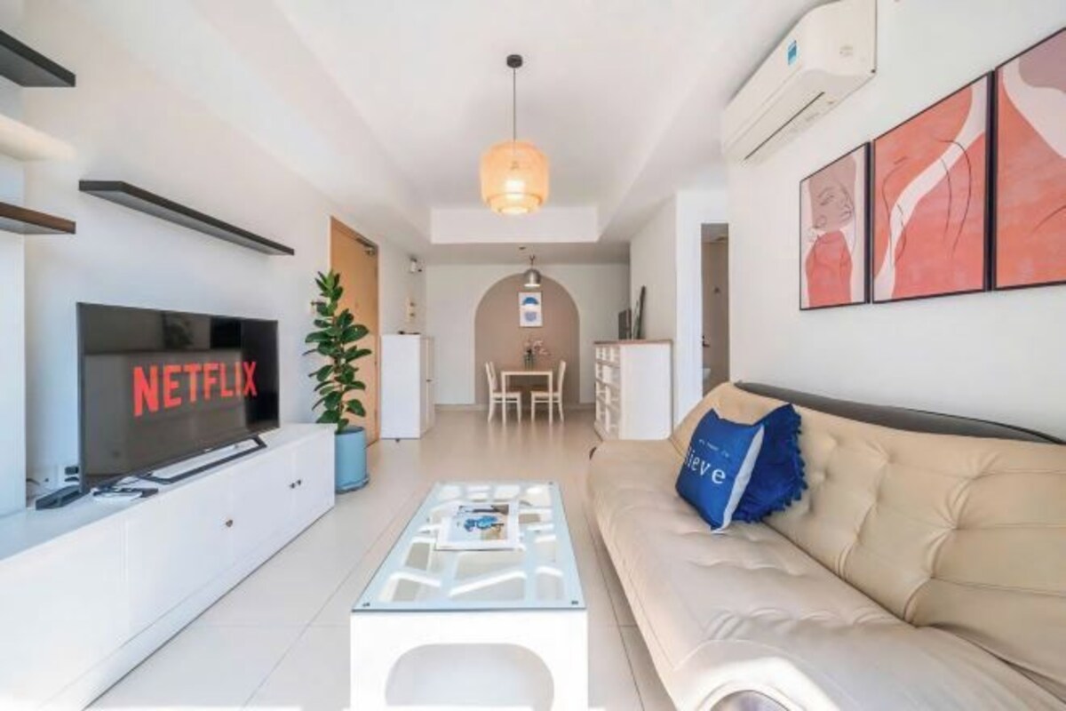 A modern living room with a flat-screen TV, a glass coffee table, a beige sofa, and a dining area in the background.