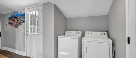 Un lavadero con dos lavadoras y secadoras blancas, una puerta con panel de vidrio y un cuadro de paisaje urbano en la pared.