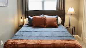 Una cama bien hecha con colcha azul, manta marrón y varios almohadones en tonos azules y marrones. Hay una lámpara de noche sobre una mesita junto a la cama.