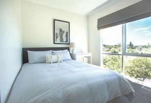 A bedroom with a large bed, a nightstand, a lamp, and a window with a view of greenery.
