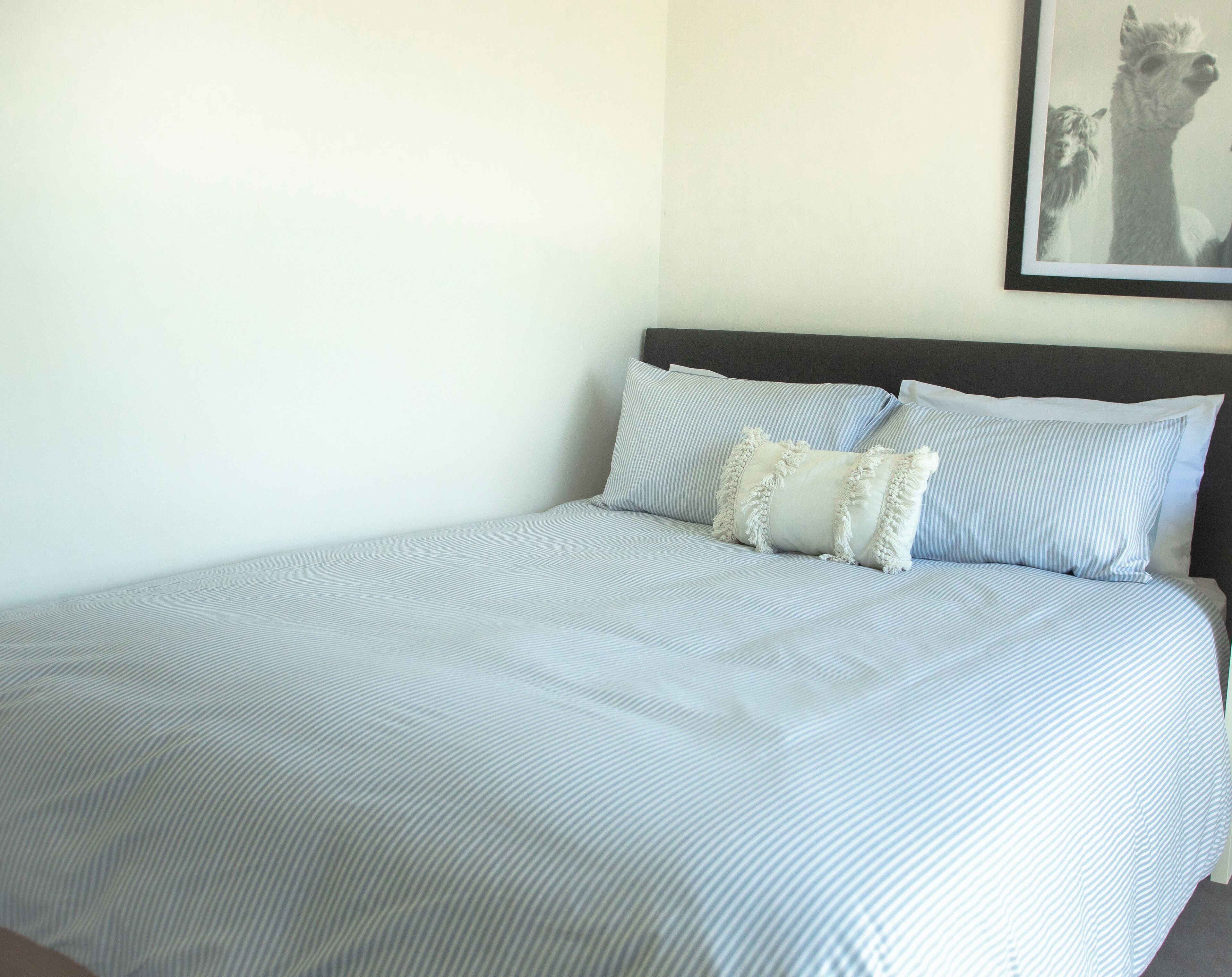 A neatly made bed with striped bedding and two pillows, a framed picture on the wall, and a plain white wall.