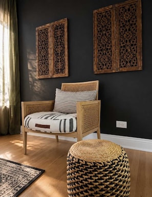 A room with a dark wall, a wooden chair with a woven backrest, a striped pillow, and a woven ottoman.