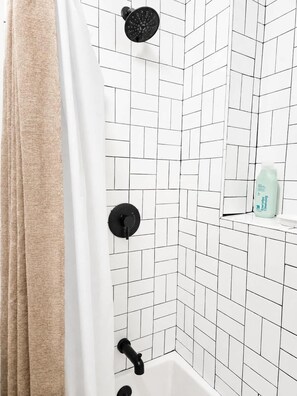 A bathroom with white tiled walls, a black showerhead, a black faucet, and a beige curtain.