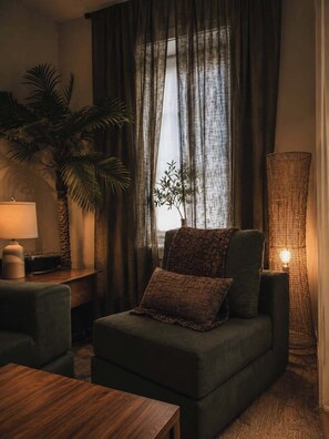 A cozy living room with a sofa, a side table with a lamp, a potted plant, and a large window with curtains. - Desert Mirage Private Patio Desk Parking (Chicago)
