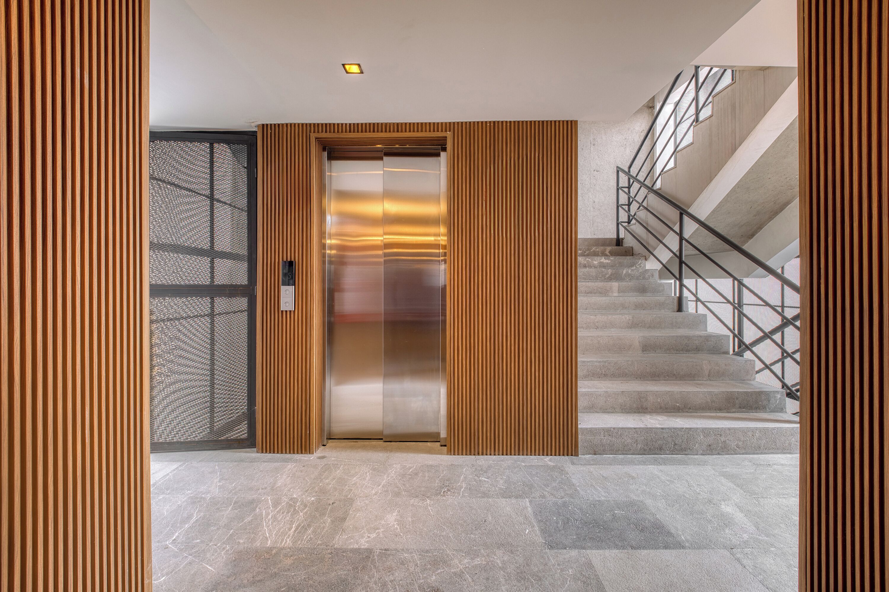 A modern elevator with a metallic finish, flanked by wooden paneling, leading to a staircase with metal railings and a concrete floor.