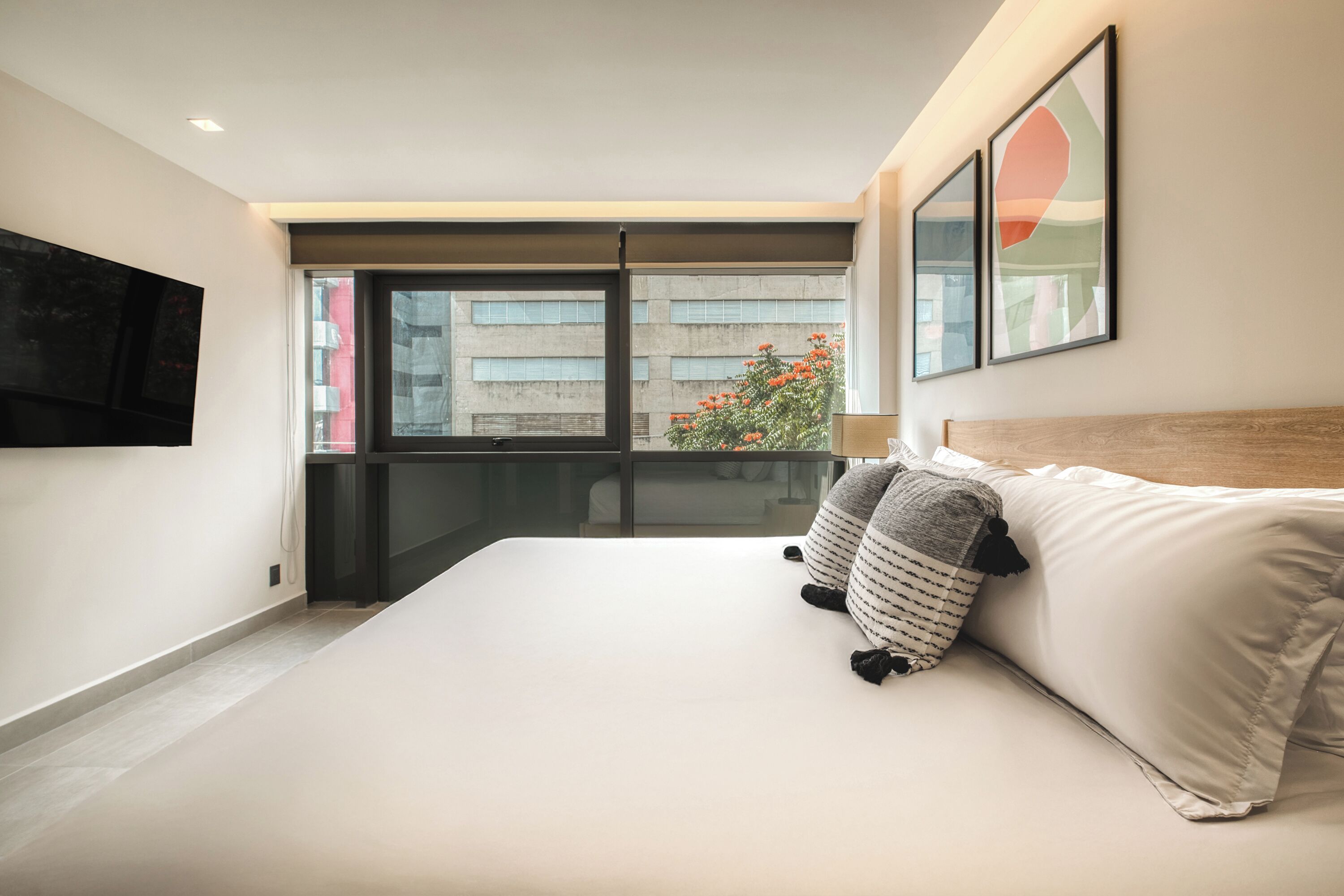 A modern bedroom with a large bed, a TV mounted on the wall, and a balcony with a city view.