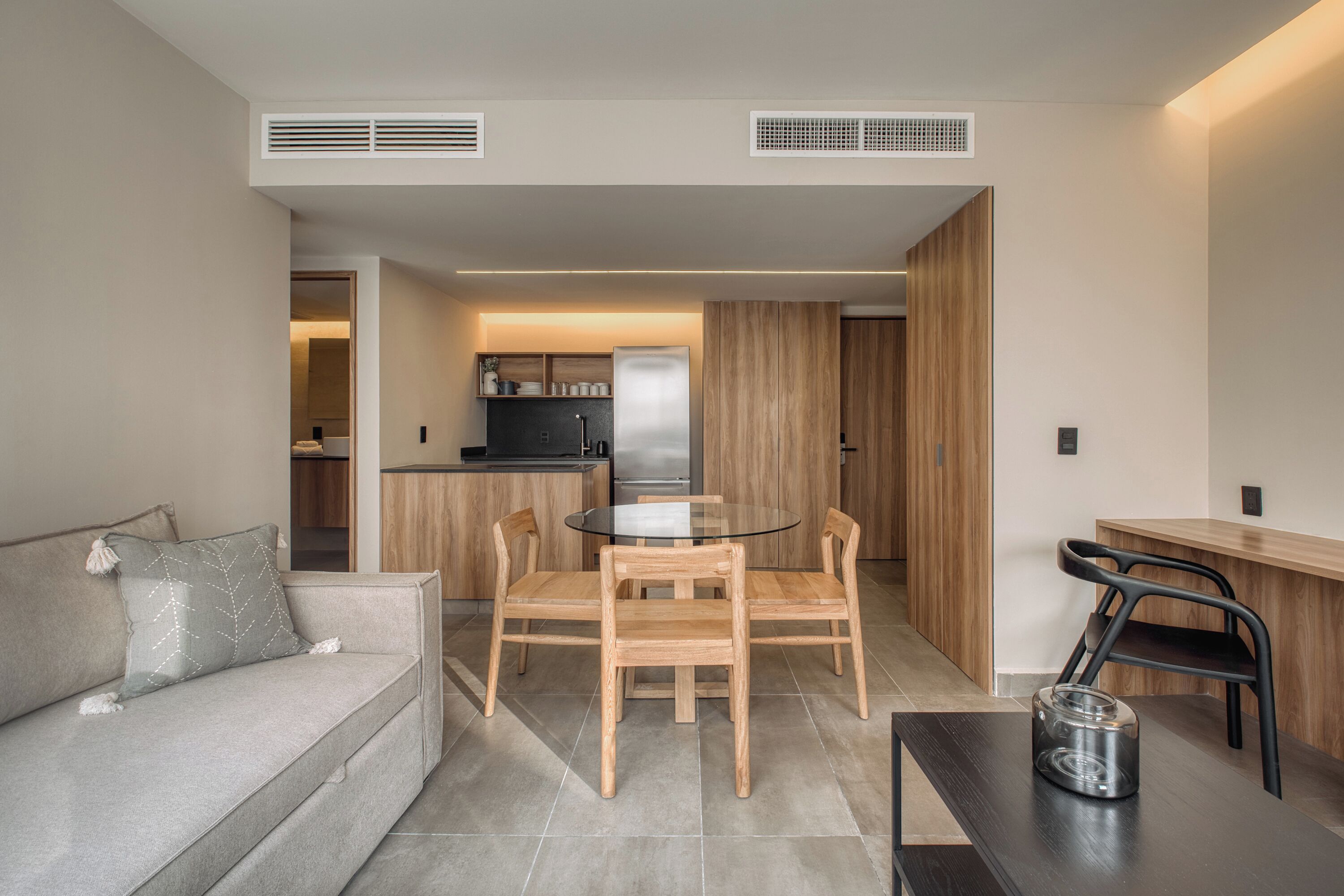 Een moderne woonkamer met een bank, een rond glazen bijzettafeltje en houten stoelen. Er is een keuken te zien op de achtergrond, met een koelkast en kastjes.
