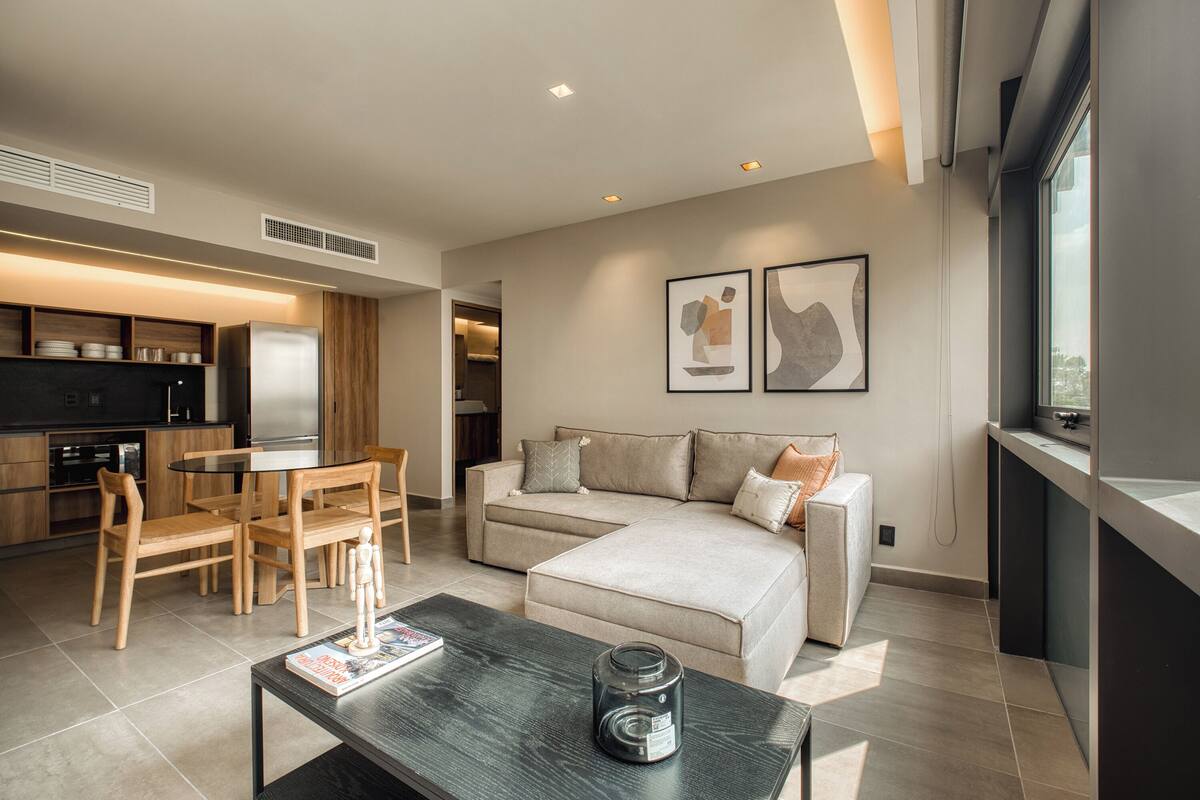 Una sala de estar moderna con sofá, mesa de centro y comedor. Se observa una cocina al fondo con refrigerador y armarios.