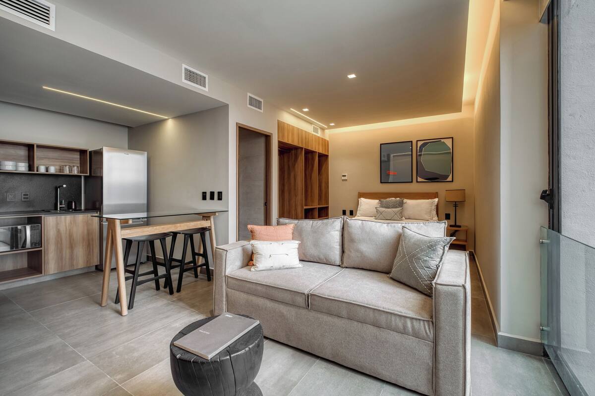 Un apartamento moderno con kitchenette, un sofá con cojines, una mesa de comedor y barandilla de vidrio.