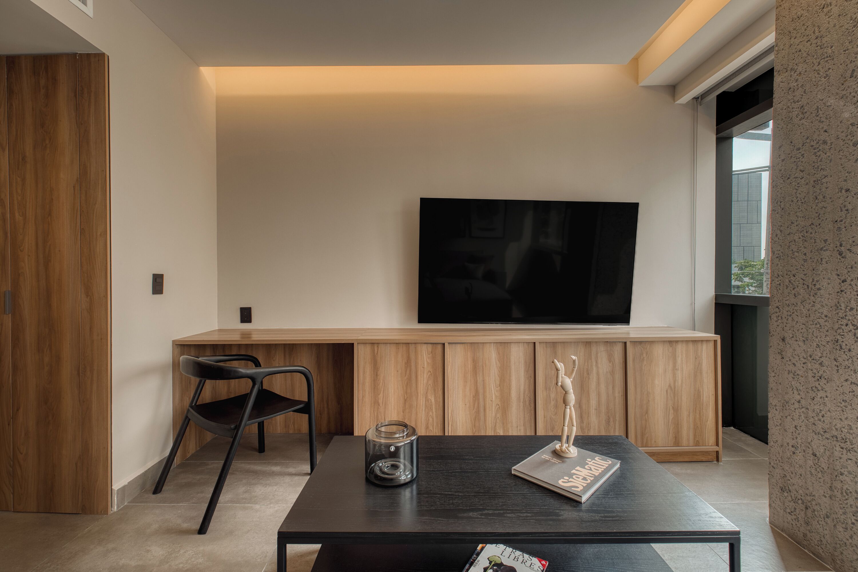 A modern living room with a flat-screen TV mounted on the wall, a wooden cabinet below, a black coffee table with a magazine and a candle, and a black chair.