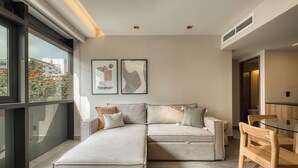 Een moderne woonkamer met een modulaire bank, een glazen eettafel en houten stoelen. Twee ingelijste kunstwerken hangen aan de muur en er is een raam met uitzicht op gebouwen en bomen.
