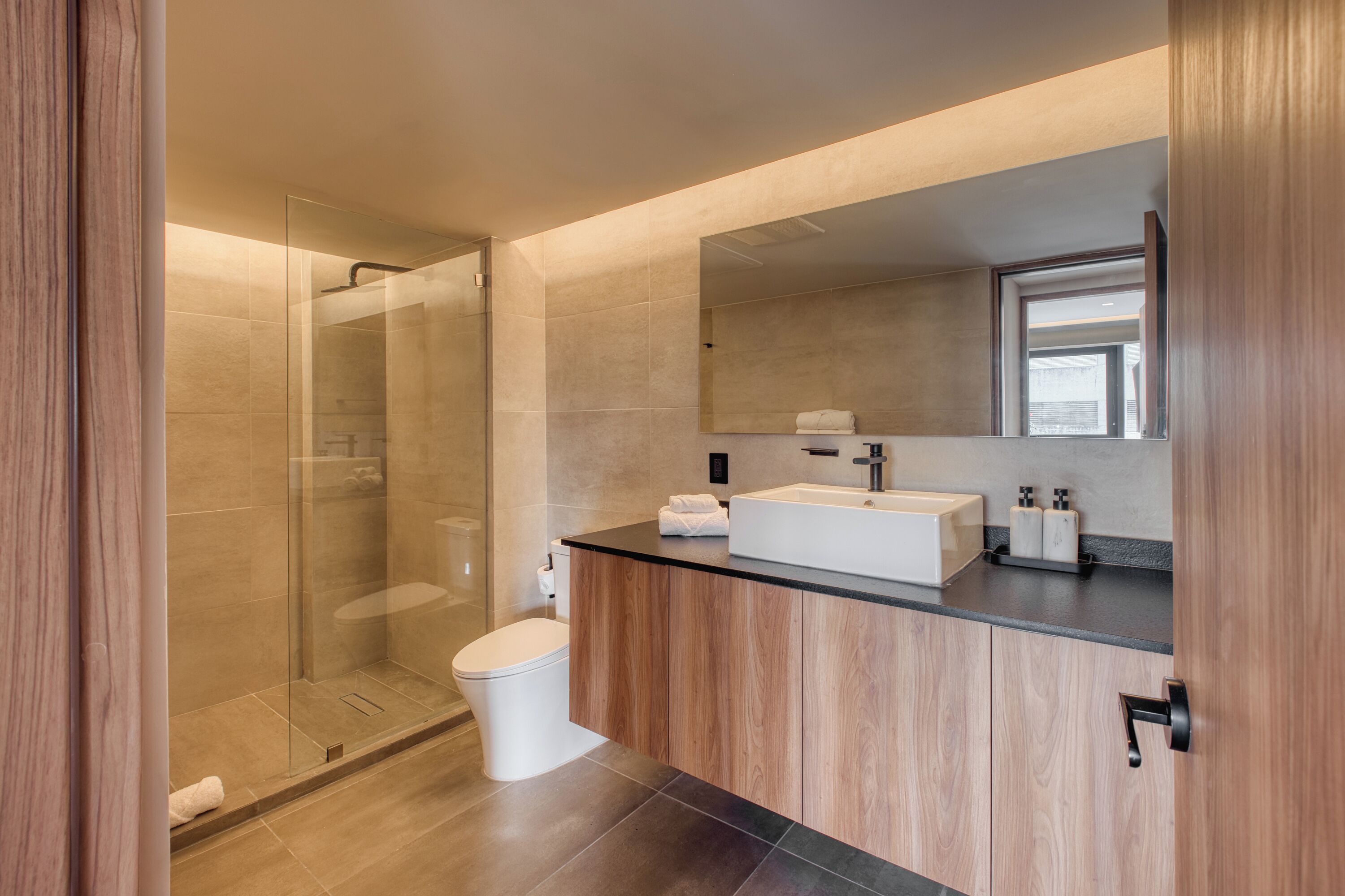 Un baño moderno con cabina de ducha de vidrio, inodoro blanco, tocador de madera con encimera negra y un espejo grande sobre el lavabo.
