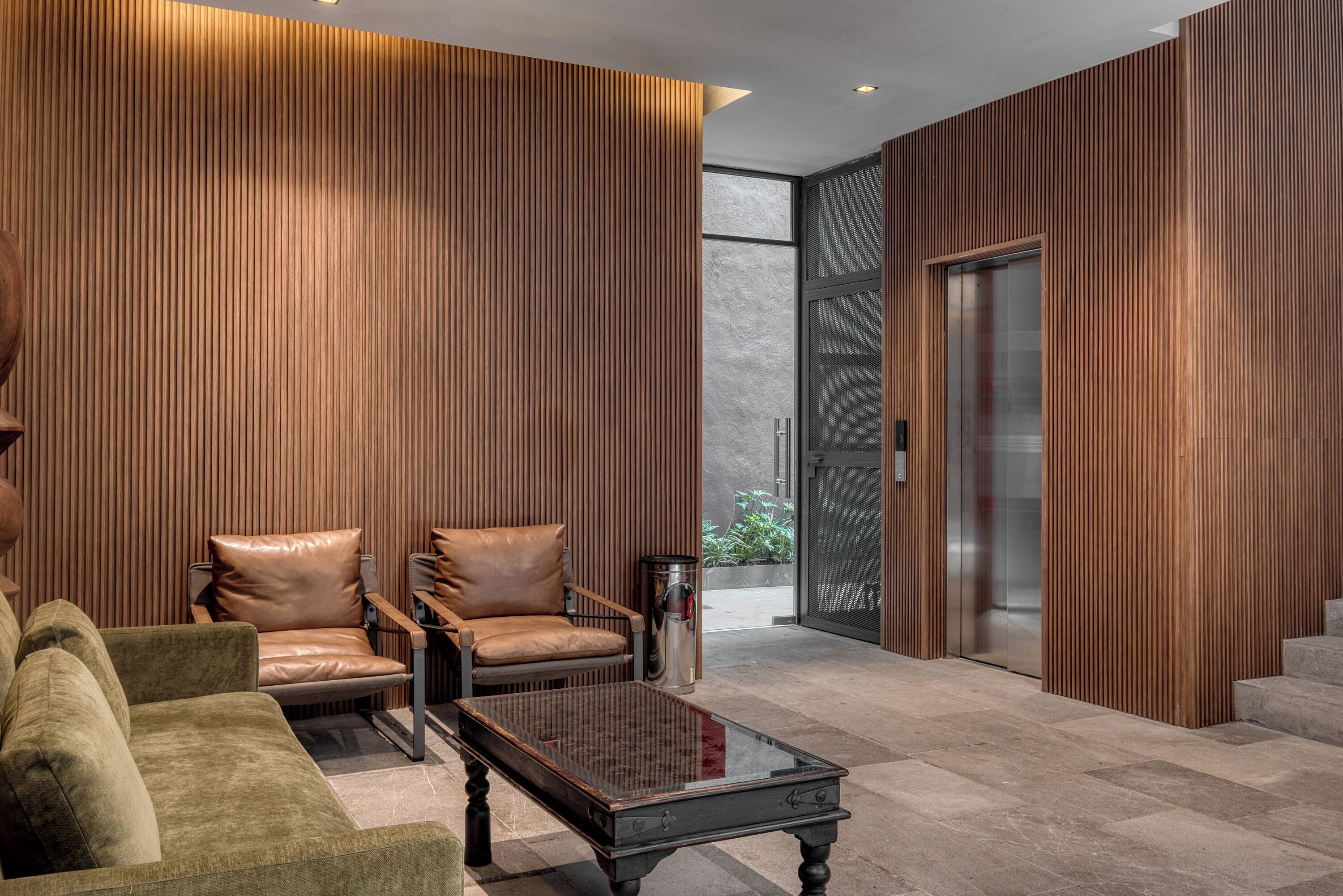 Een moderne lobby met een houten paneelwand, lederen stoelen, een glazen salontafel en een glazen deuropening van een lift.
