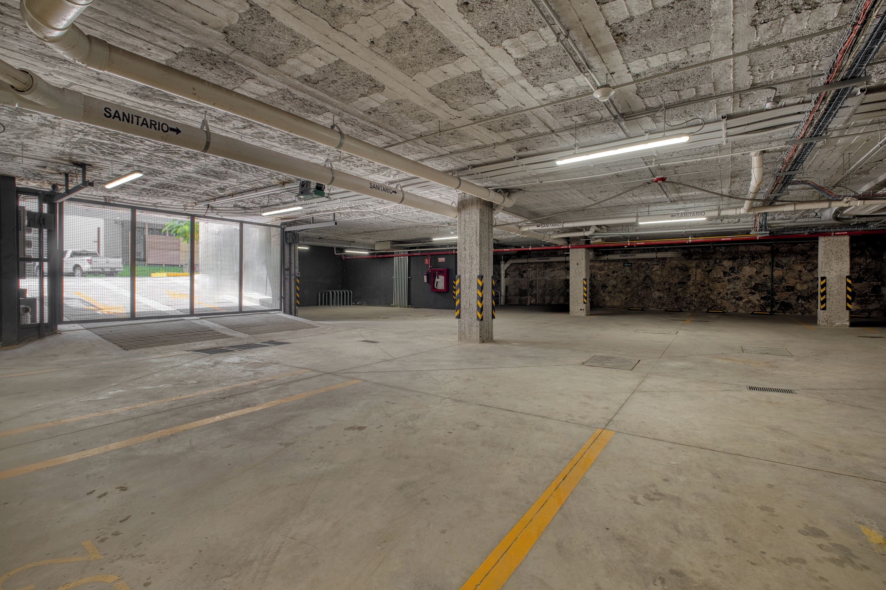 Garagem subterrânea com pilares de concreto, linhas amarelas no chão e placas indicando 'SANITÁRIO'.