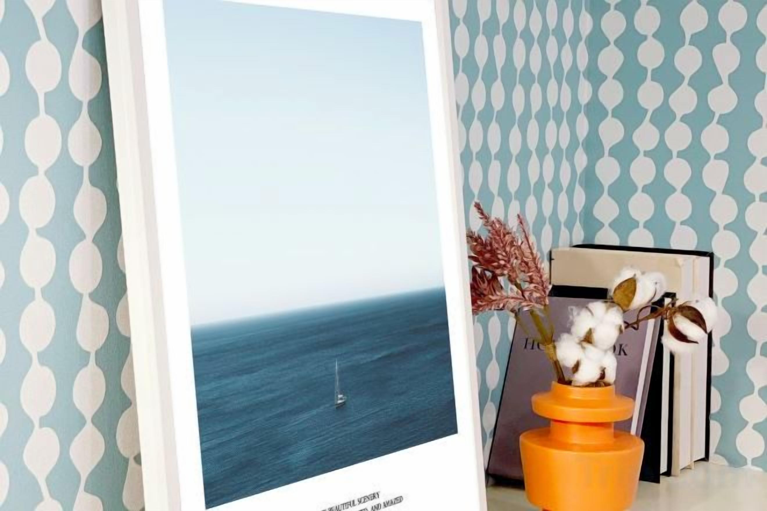 A framed photograph of a calm sea with a sailboat, a vase with dried plants, and books on a shelf against a patterned wall.