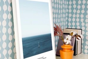 Une photographie encadrée représentant une mer calme avec un voilier, un vase contenant des plantes séchées et des livres sur une étagère, le tout placé devant un mur à motifs.