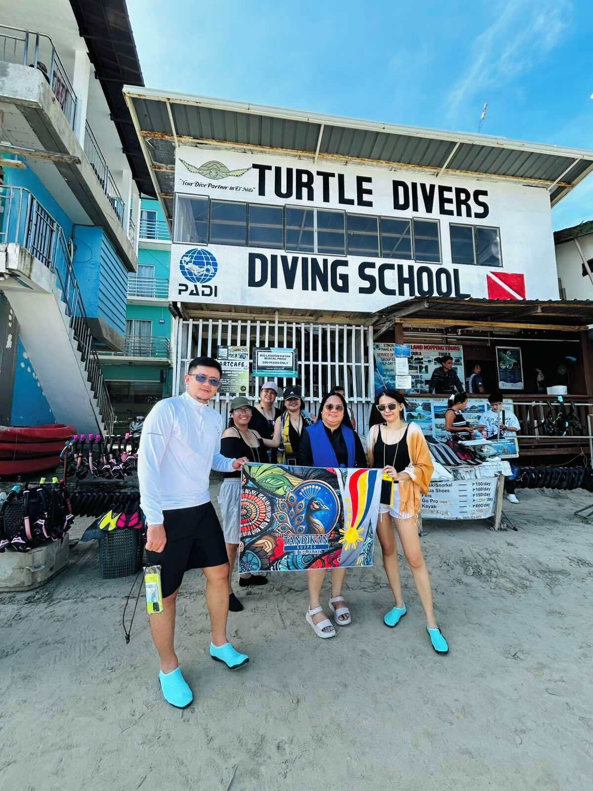 Un grupo de personas frente a un edificio con la leyenda 'Turtle Divers Diving School', un letrero de 'PADI' y un banderín que dice 'TANDIKAN'.