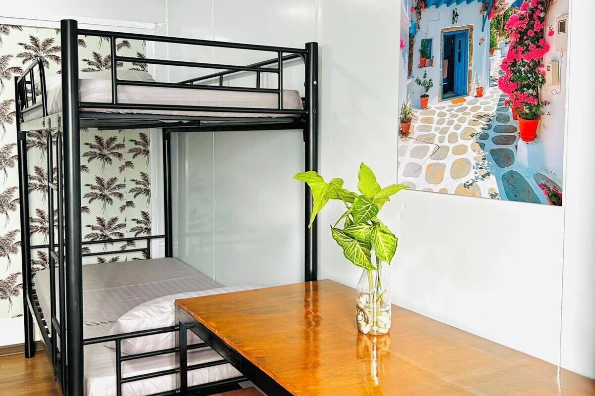 Una cama individual con una mesita de madera y una planta en maceta en una habitación con paredes blancas y un mural azul y blanco.