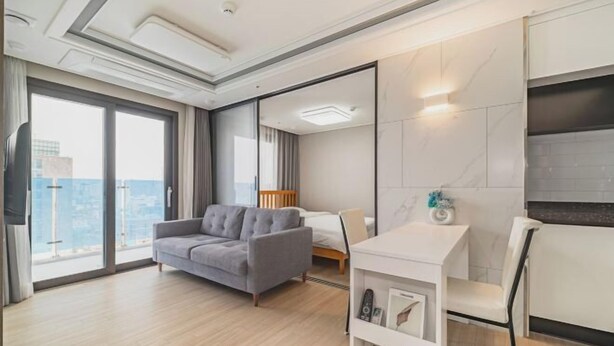 Une chambre d’hôtel moderne avec une grande fenêtre, un canapé gris, un lit, une petite table blanche et un téléviseur.