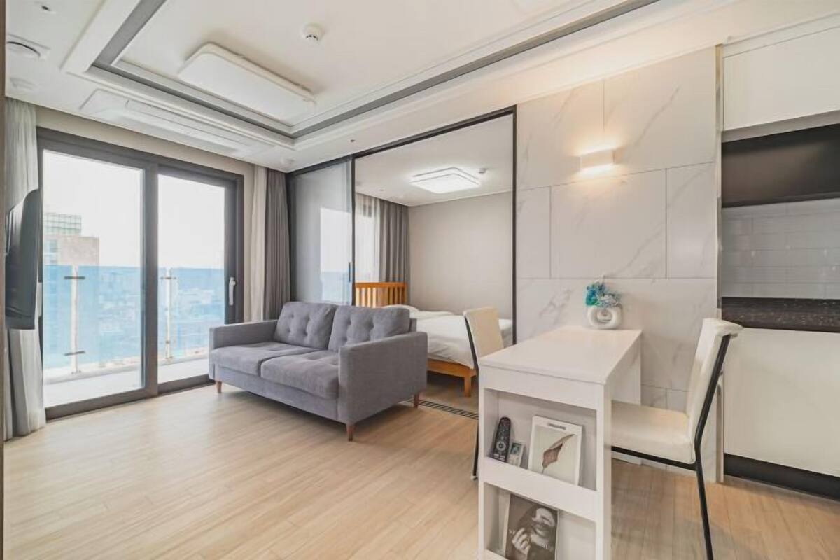Une chambre d’hôtel moderne avec une grande fenêtre, un canapé gris, un lit, une petite table blanche et un téléviseur.