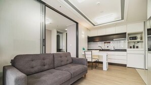 Un salon moderne avec un canapé gris, une table à manger avec des chaises et un coin cuisine doté de meubles blancs et d’un four à micro-ondes.