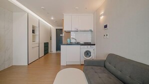 Un appartement moderne doté d’une kitchenette, d’un lave-linge, d’un canapé gris et d’un pouf blanc.