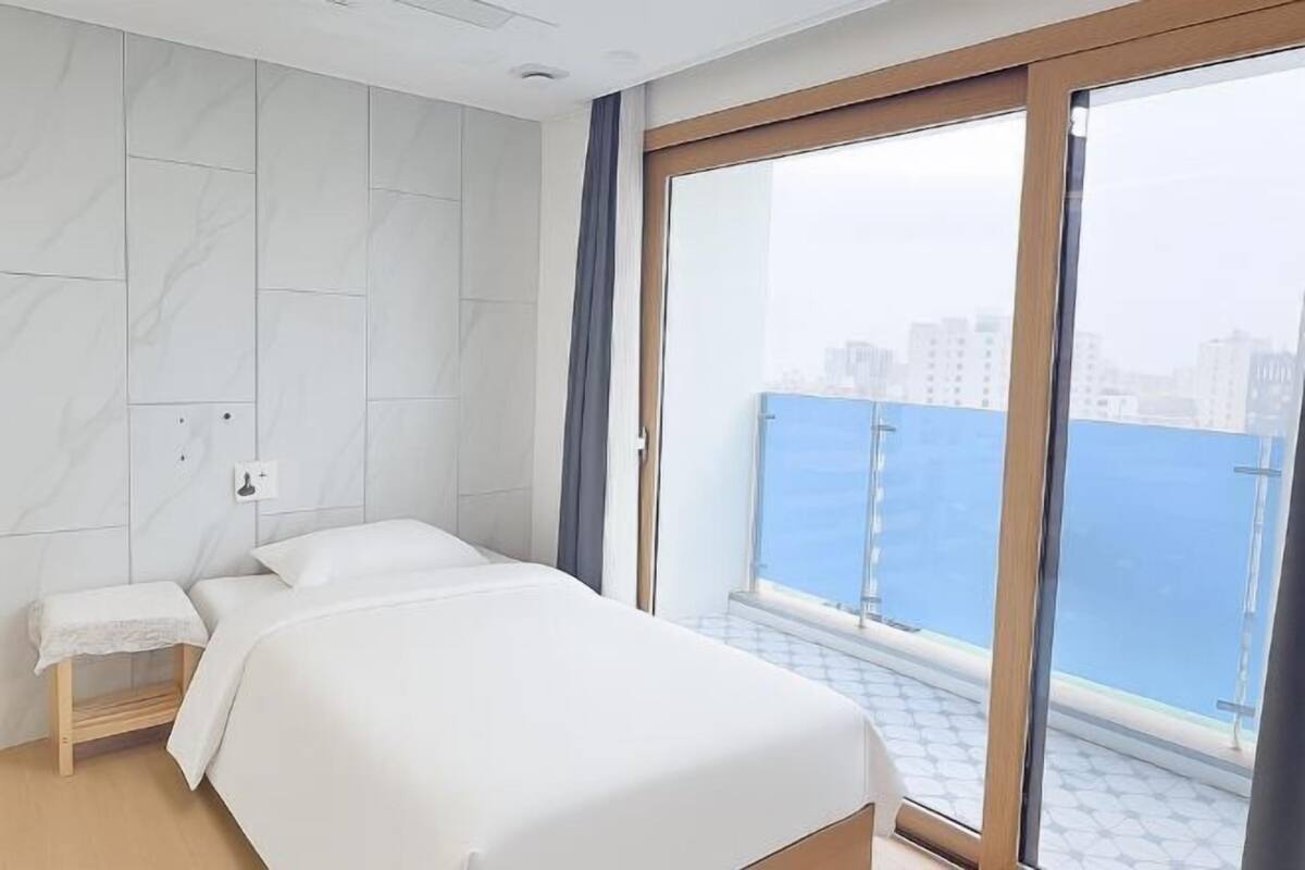 Une chambre d’hôtel moderne, dotée d’un grand lit, d’une petite table de chevet et offrant une vue sur la mer grâce à une porte-fenêtre coulissante.